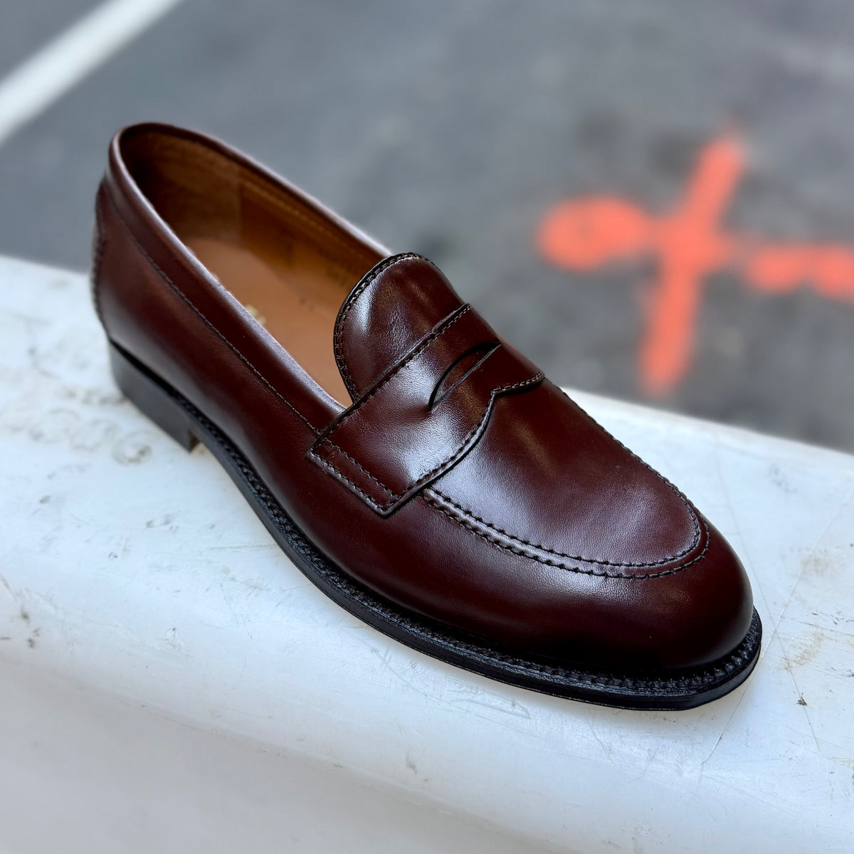 A single D5214 - Brown Calf Flex Penny by Alden Madison, elegantly crafted on the Copley Last in rich Brown Vegano Calf leather, sits on a white surface with a blurred street and orange marking in the background. Its Flex Leather Sole ensures comfort while complementing the loafer’s timeless silhouette.