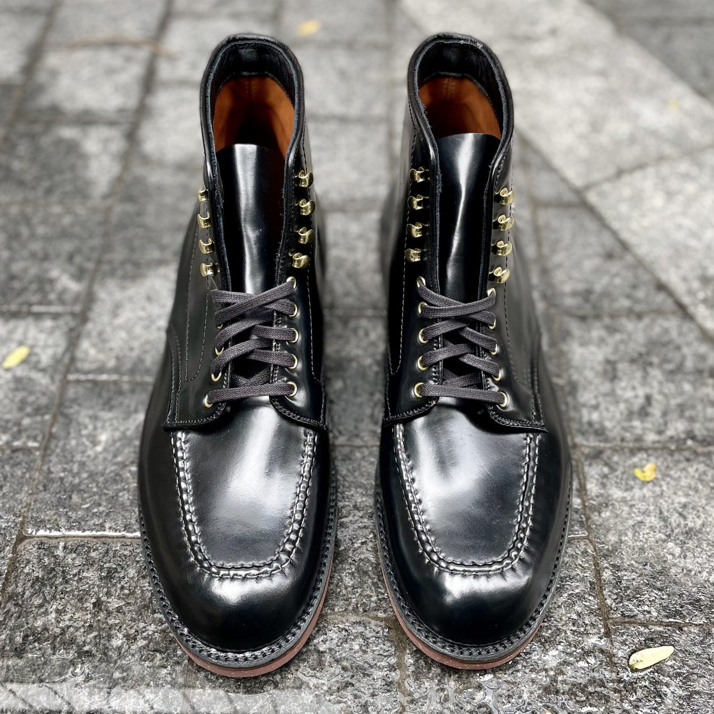 The Alden Madison D1908H Copake Indy Boot in Black Shell Cordovan features detailed hand stitching and gold eyelets, displayed side by side on a textured stone pavement.