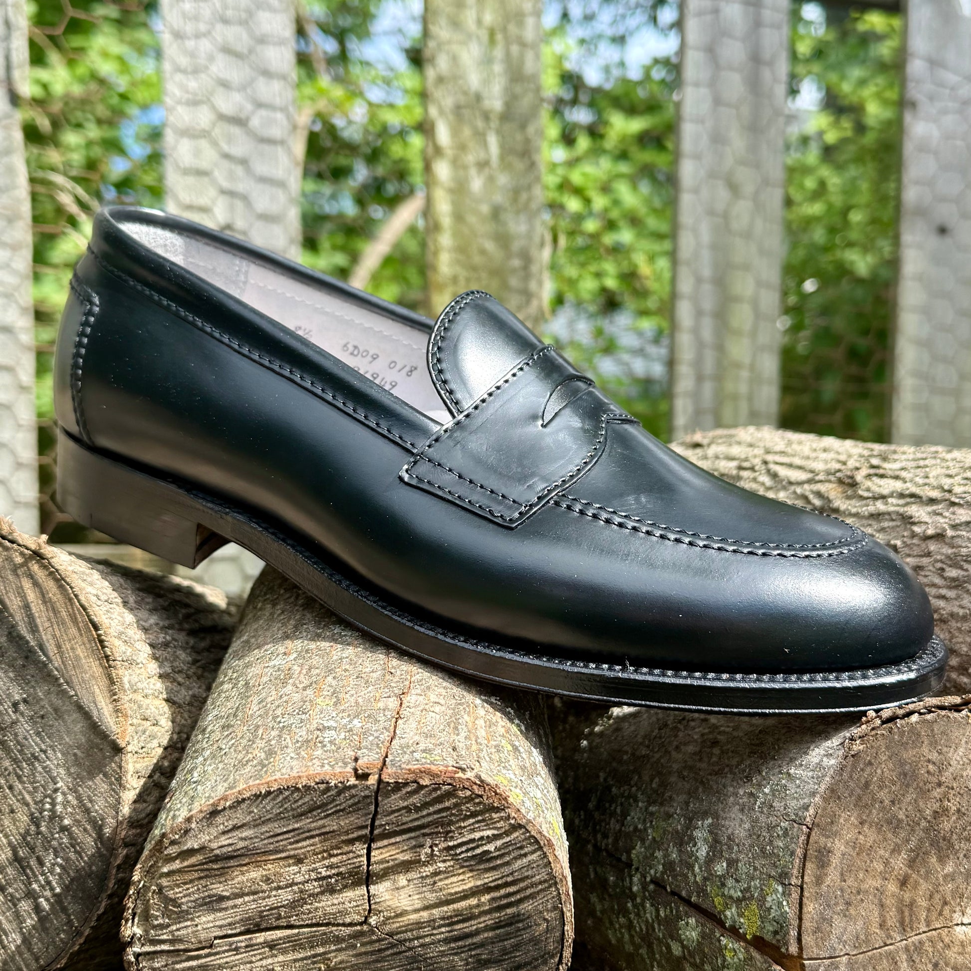 The Alden Madison 96949 - Copley Penny Loafer in Black Shell Cordovan, crafted on the Copley Last with a refined 270 Flat Welt, is displayed outdoors on stacked logs, set against a blurred backdrop of wooden rails and green foliage.