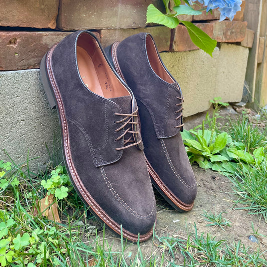 A pair of Alden Madison 704 Mocc Toe Bluchers in brown suede with a dress rubber sole rest on the ground near grass and plants, with a brick wall and blue flowers in the background.