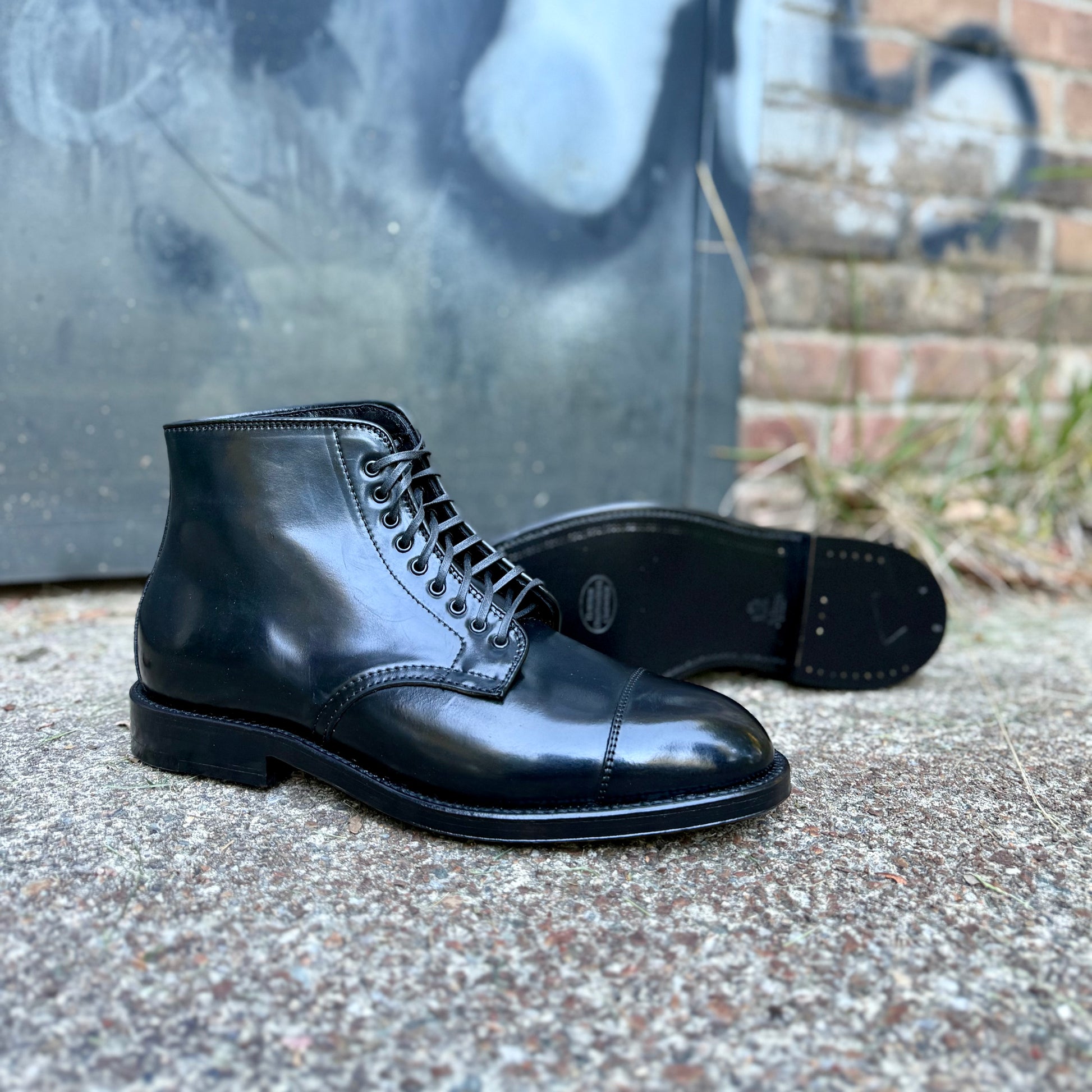 The Alden Madison 4065 Cap Toe Boot in Black Shell Cordovan, featuring a Double Oak Leather Sole, is displayed outdoors on rough concrete with one boot upright and one on its side; a brick wall and graffiti-covered metal door are in the background.