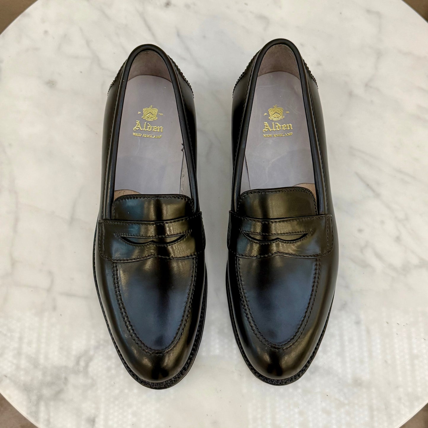 A pair of ALDEN SHOES 5751 High Vamp Loafers in black shell cordovan, crafted on the Aberdeen Last, rest side by side on white marble. Gold Alden logos and text are stamped on the insoles.
