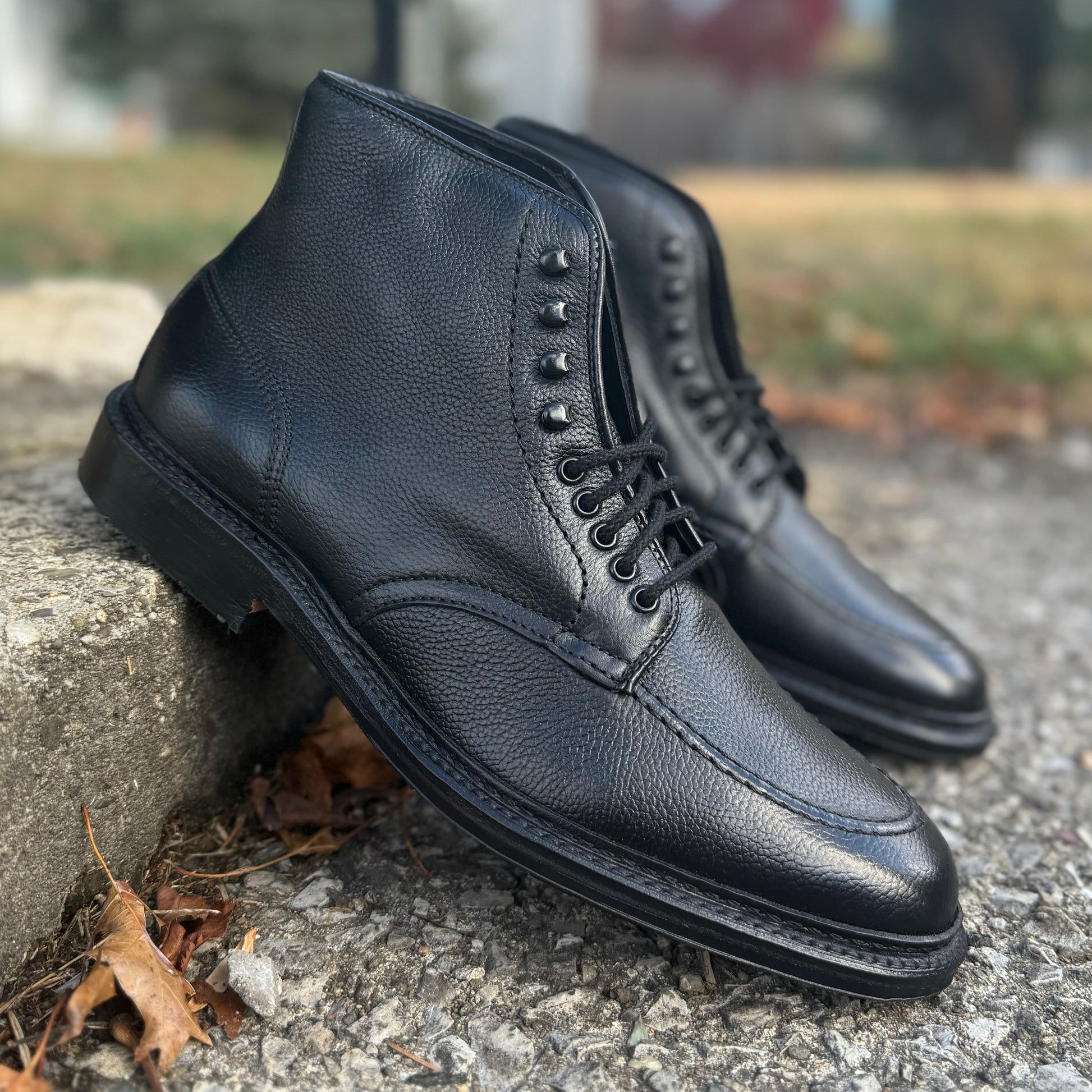 Alden Madison’s D6956LH "Han" Boot in Black JS Grain leather, inspired by the Indy Boot style, features textured uppers and is crafted on the Barrie Last for comfort and durability. Shown outdoors on rough concrete with dry leaves nearby.
