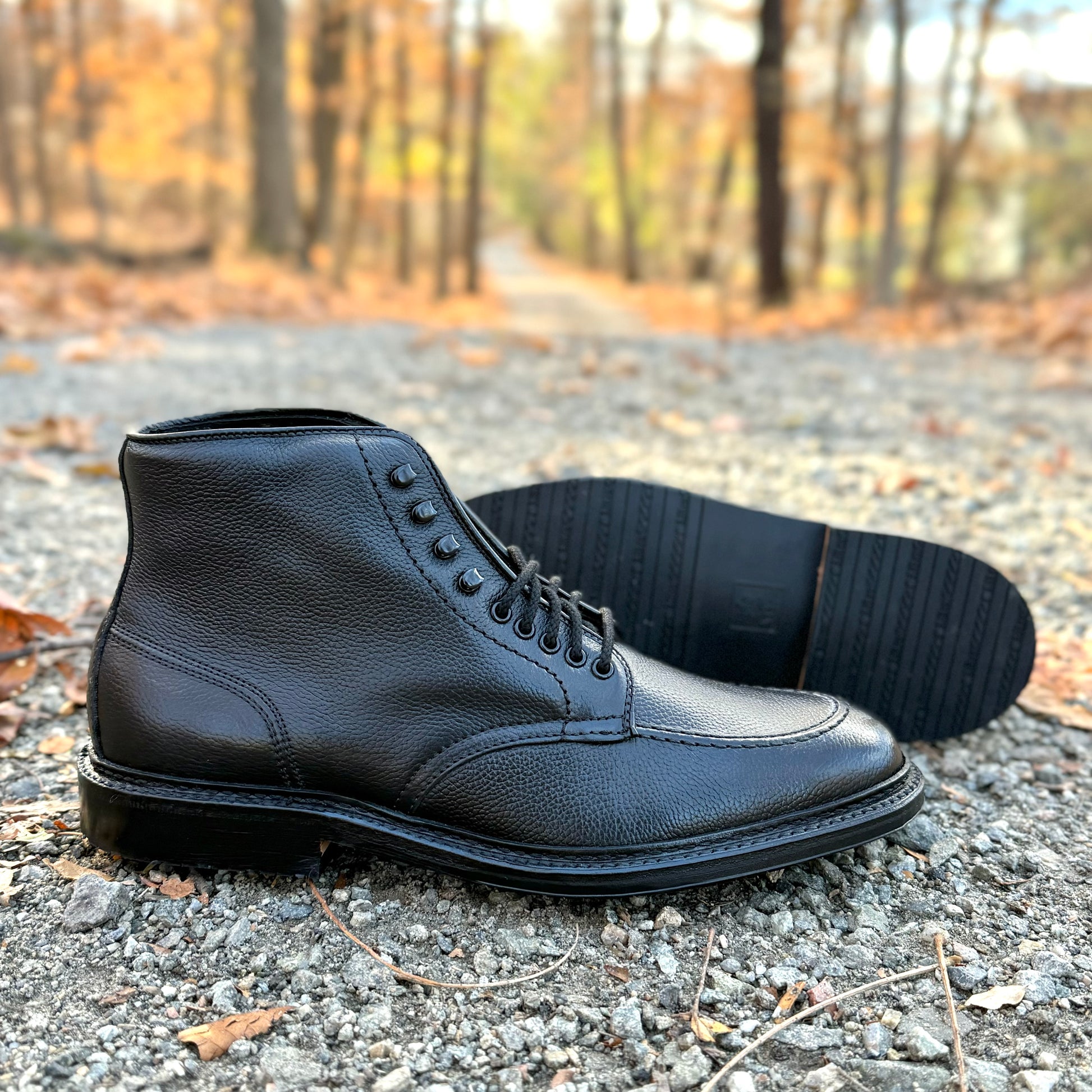 The Alden Madison D6956LH "Han" Boot in Black JS Grain, with one upright and one on its side, is shown on a gravel forest path among autumn leaves. Crafted on the Barrie Last for classic style and comfort.