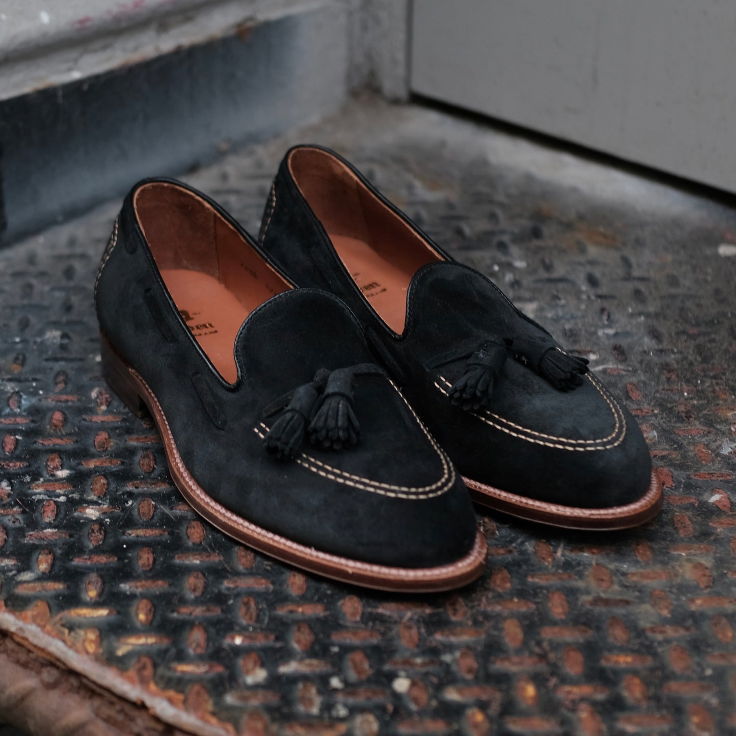 The Alden Madison D0106 Black Suede Tassel Loafers, featuring brown leather soles and white stitching, are displayed on a textured metal surface near a gray door.