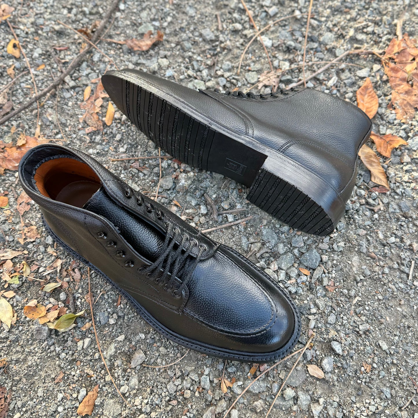Two Alden Madison "Han" Boots (D6956LH) in Black JS Grain leather rest outdoors on gravel with dry leaves. One upright on the Barrie Last, the other on its side shows off the rugged sole.