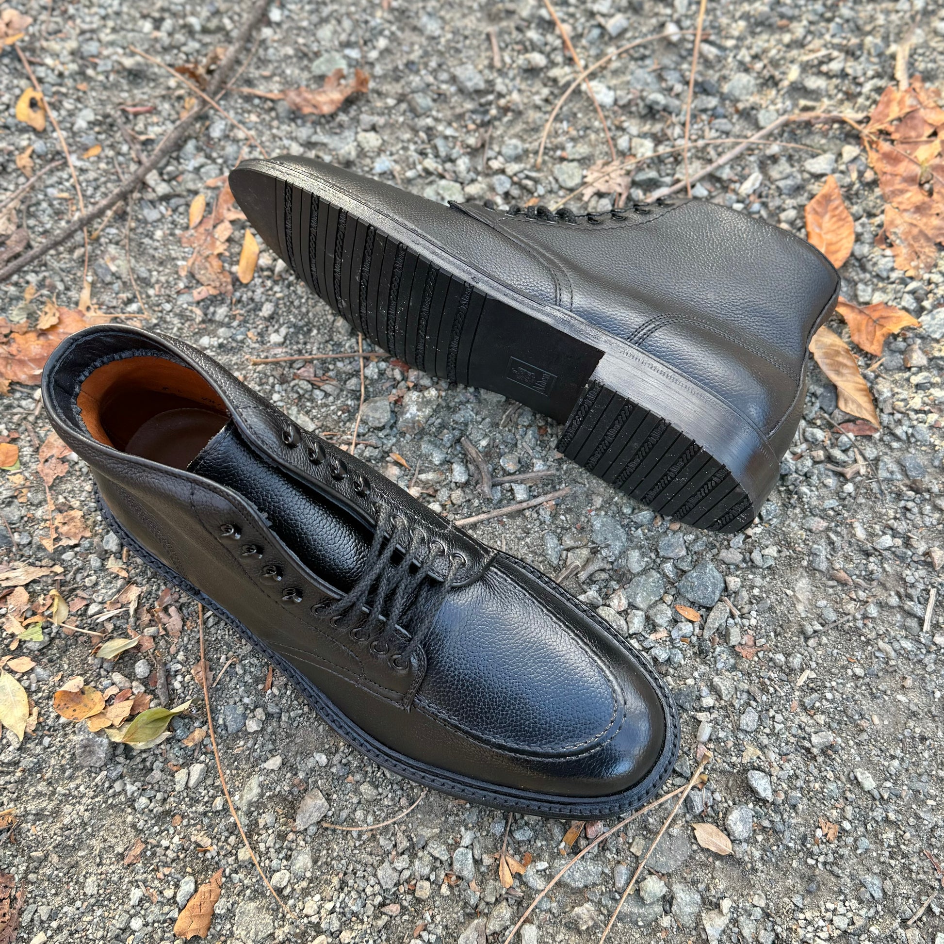 Two Alden Madison "Han" Boots (D6956LH) in Black JS Grain leather rest outdoors on gravel with dry leaves. One upright on the Barrie Last, the other on its side shows off the rugged sole.