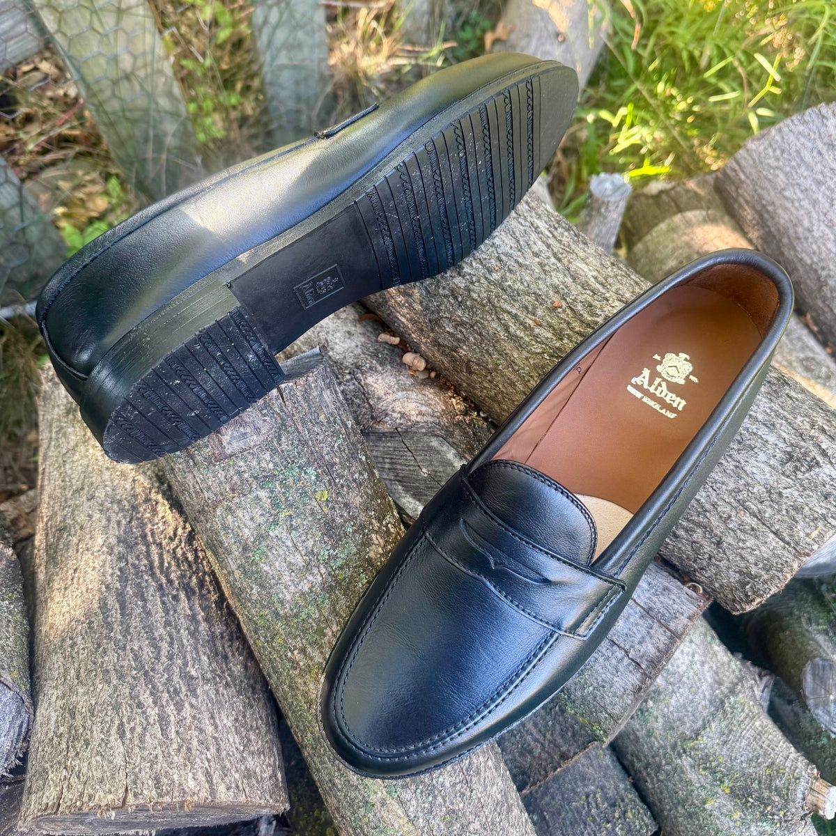 A pair of ALDEN SHOES D5219L Penny Loafers in Black Lady Calf leather, featuring a sturdy rubber sole, are displayed outdoors on stacked tree logs—one shoe standing upright and the other revealing the sole.