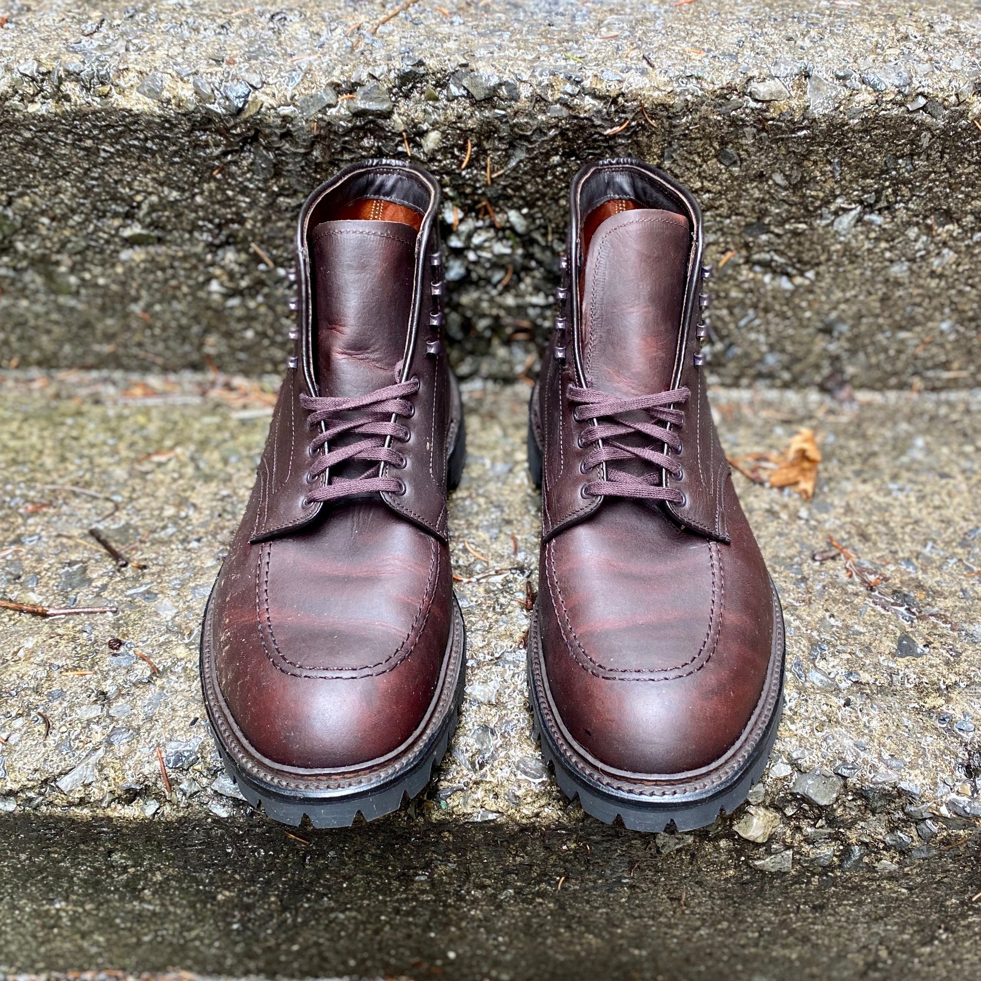 A pair of Alden Madisons 404 - Indy Boot in Kudu (Oiled CXL) rests on concrete steps. Featuring rugged kudu leather, Vibram lug outsoles, and metal eyelets, they blend durability with style. The steps beneath showcase a rough, weathered look.