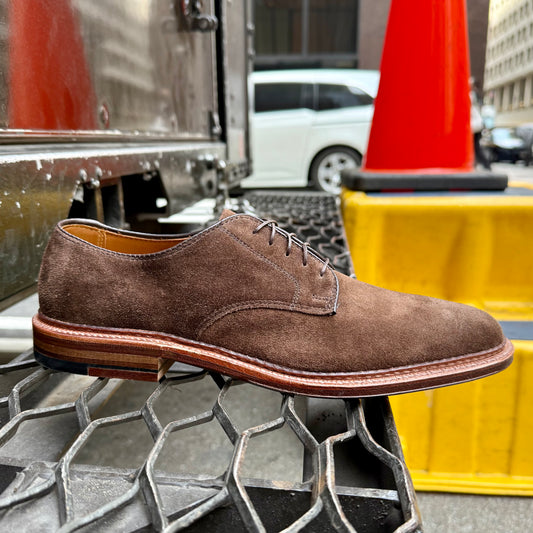 The Alden Madison 29335F - Unlined Dover in Brown Suede, crafted on the Barrie Last with a Flex Leather Sole, is displayed on a metal grate in an urban scene featuring a traffic cone and yellow barrier in the background.