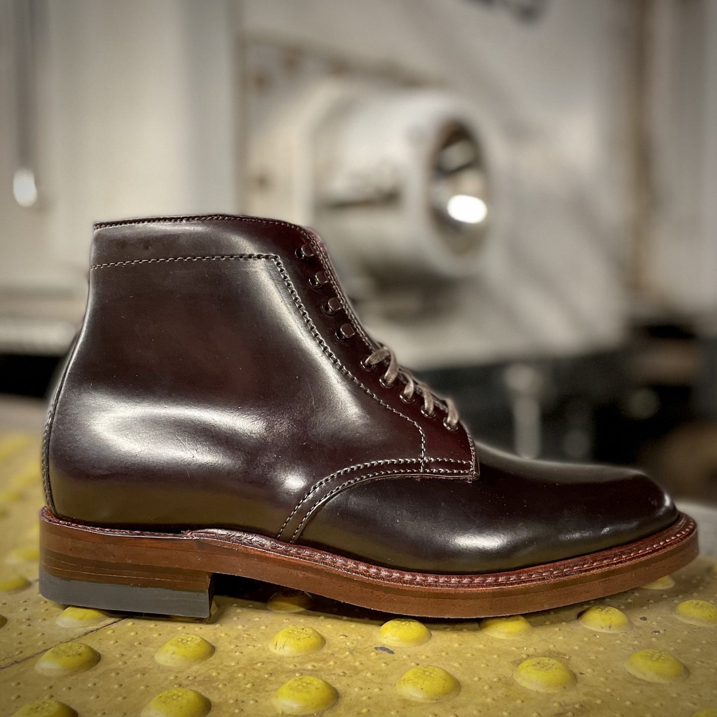 The Alden Madison D1854H - Plain Toe Boot w Neocork Sole in Color 8 Shell Cordovan, featuring a stacked heel and a rich dark brown finish, sits on a yellow textured surface against a blurred industrial backdrop.