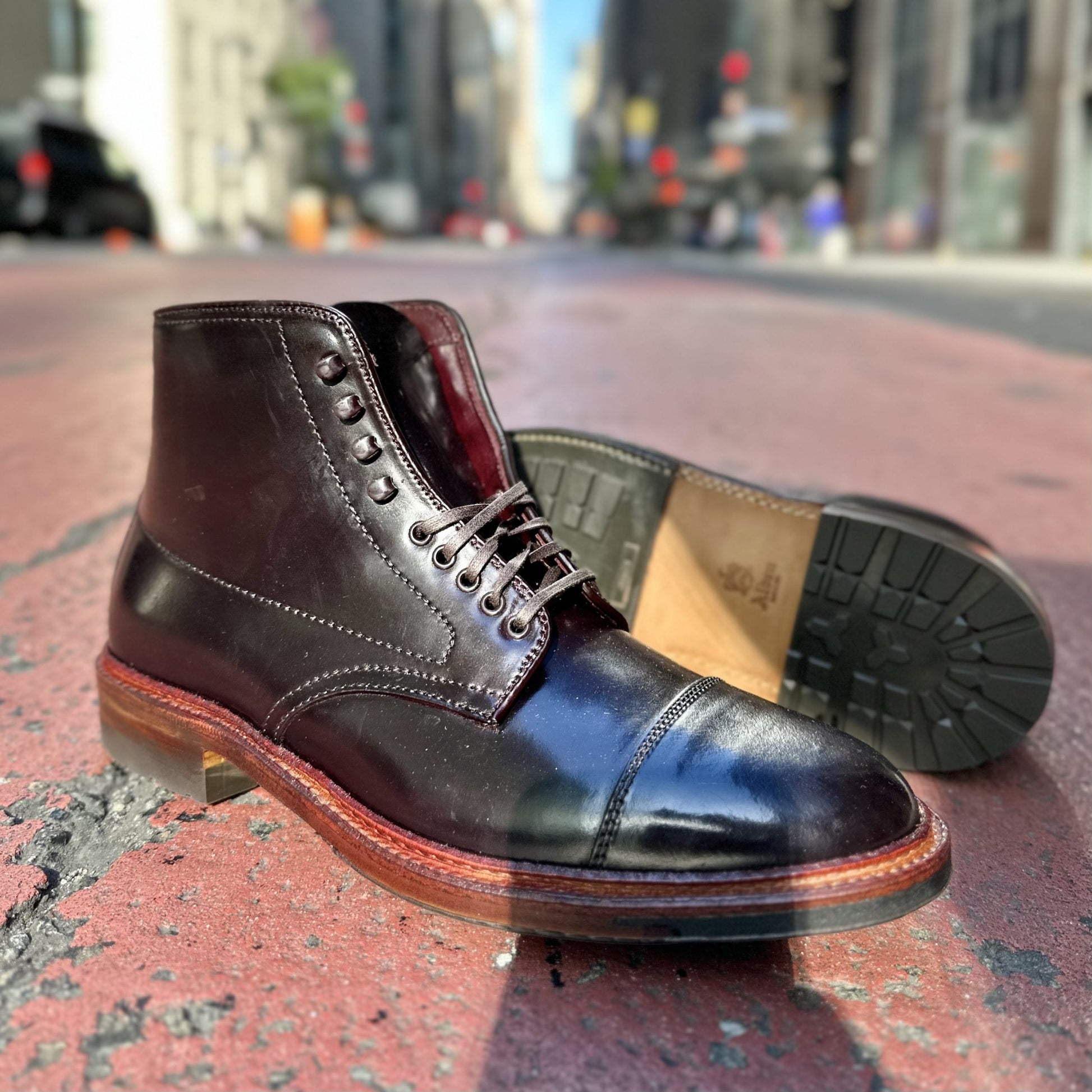 The Alden Madison D2824HC - Cap Toe Boot in Color 8 Shell, featuring a sturdy Commando sole, is displayed on an urban street. One boot stands while the other lies on its side, exposing the robust sole against a backdrop of blurred city buildings and cars.