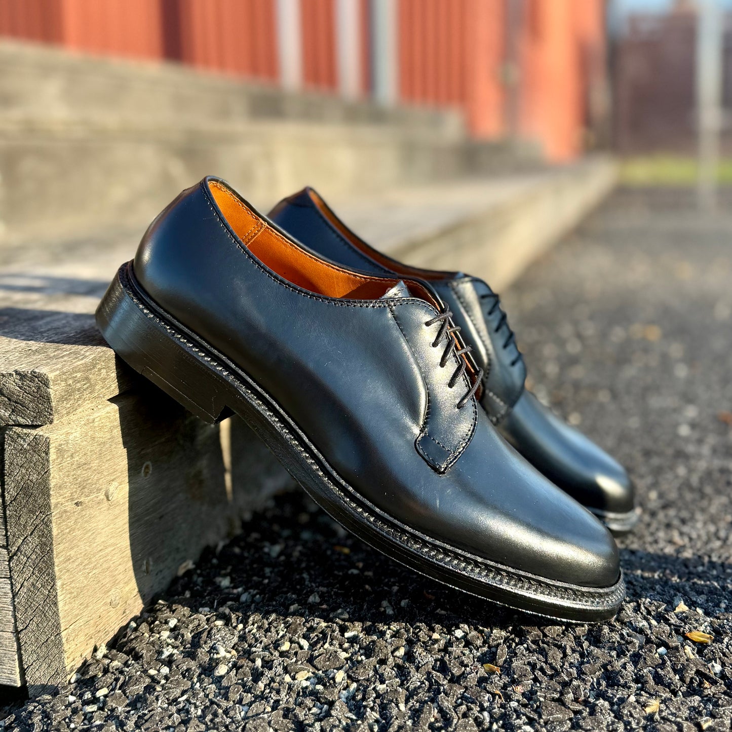 A pair of Alden Madison 9901 Plain Toe Blucher shoes in shiny black Shell Cordovan with brown lining and Double Oak Leather Sole sits on a wooden bench, blurred red building and pavement in the background.