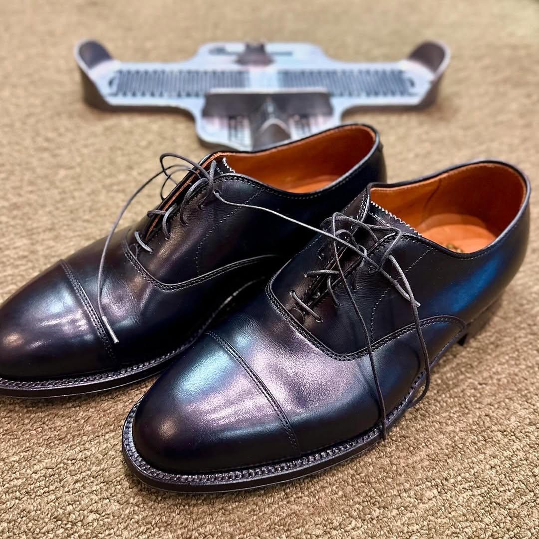 A pair of Alden Madison 907 - Straight Tip Bal dress shoes in polished black calfskin with laces rests on a beige carpet, with a metal shoe sizer visible in the background.