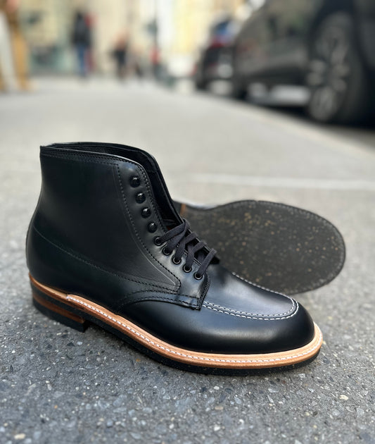 The Alden Madison 401 - Indy Boot in Black Chromexcel, featuring a tan welt and Neoprene cork sole, is shown on a city sidewalk—one boot upright and one on its side to highlight the durable sole construction.