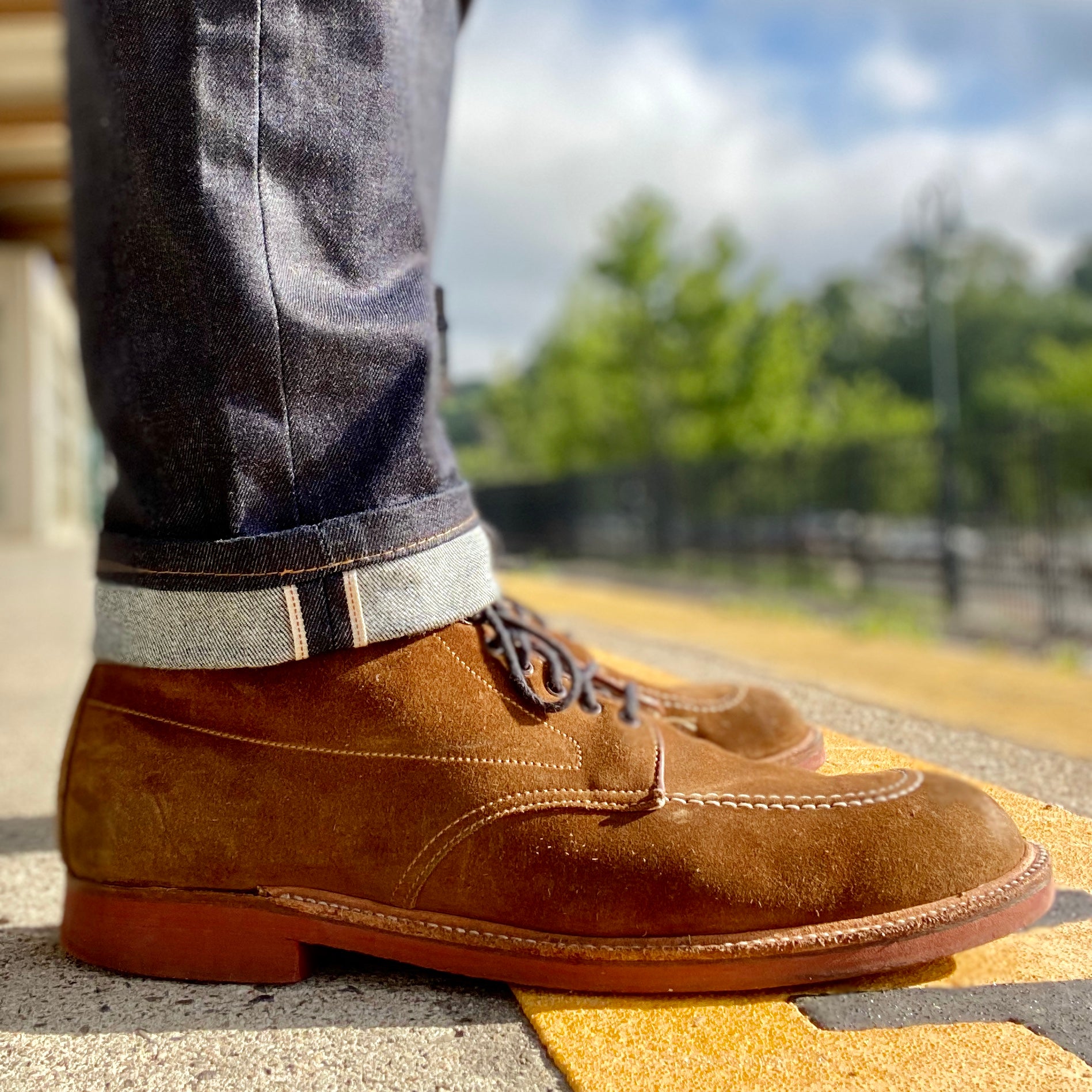 Close-up of a person wearing Alden Madisons 40531H Redwood Indy Boot in Snuff Suede, paired with dark denim jeans with rolled cuffs, standing on yellow tactile paving. The blurred background reveals greenery and a fence beneath a cloudy sky.