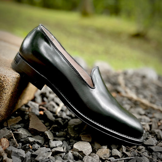 An 8815 Plain Toe Slip-On in Black Shell Cordovan by Alden Madison rests on its side among gray stones, set against a blurred green backdrop. The loafer, crafted on the Aberdeen Last, showcases a polished finish and low heel atop a raised edge.