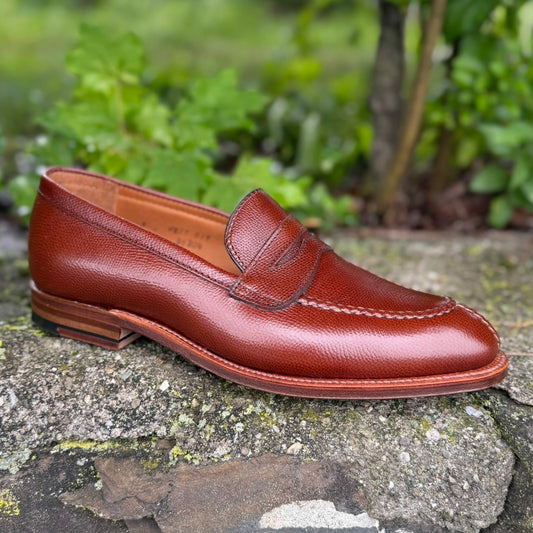 The Alden Madison D1204 - Alpine Grain NST Loafer, crafted in single brown textured grain leather with an antique flat welt, rests on a stone surface outdoors, green foliage softly blurred in the background.