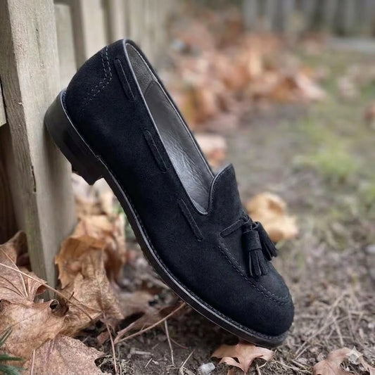 The ALDEN SHOES 36102 - Black Suede Tassel Loafer, crafted on the Aberdeen Last, rests by a wooden fence, surrounded by dry fallen leaves.