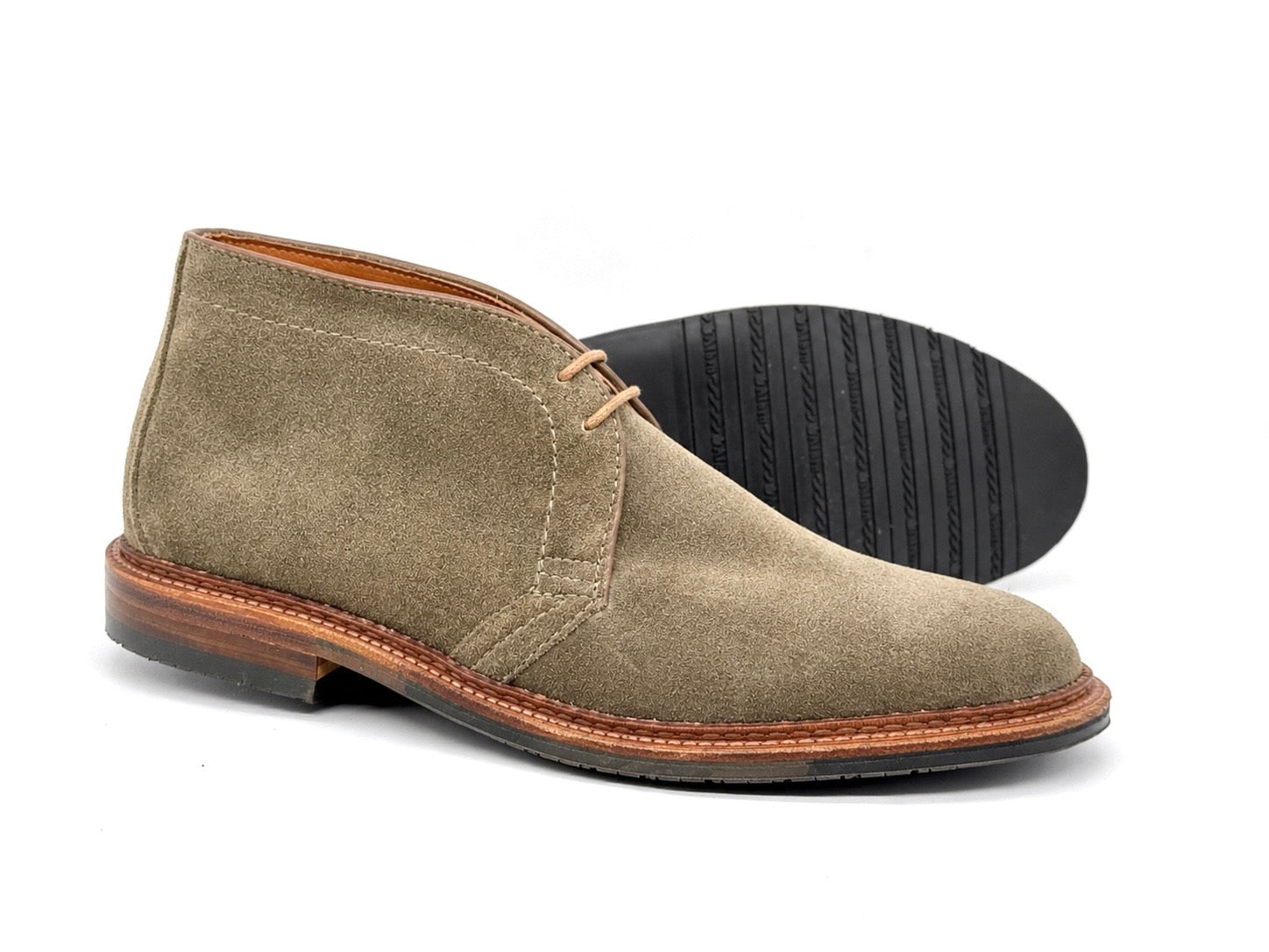 A pair of ALDEN SHOES 1590L Clay Suede Chukka Boots with brown leather soles, black rubber outsoles, and two eyelets; one shoe stands upright while the other is angled to show the sole.