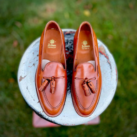 Alden Madisons 662 Tassel Loafers in burnished tan calfskin, featuring decorative stitching and a leather sole on the Aberdeen Last, are elegantly showcased on a weathered round stool outdoors against lush grass.