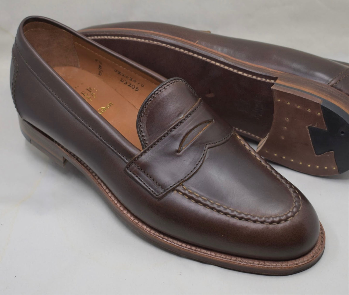 A pair of Alden Madison D5211 - Brown CXL Flex Penny Loafers on the Copley Last—one flat, one on its side—highlight the visible stitching and showcase the rich leather sole and heel detailed with brass nails.