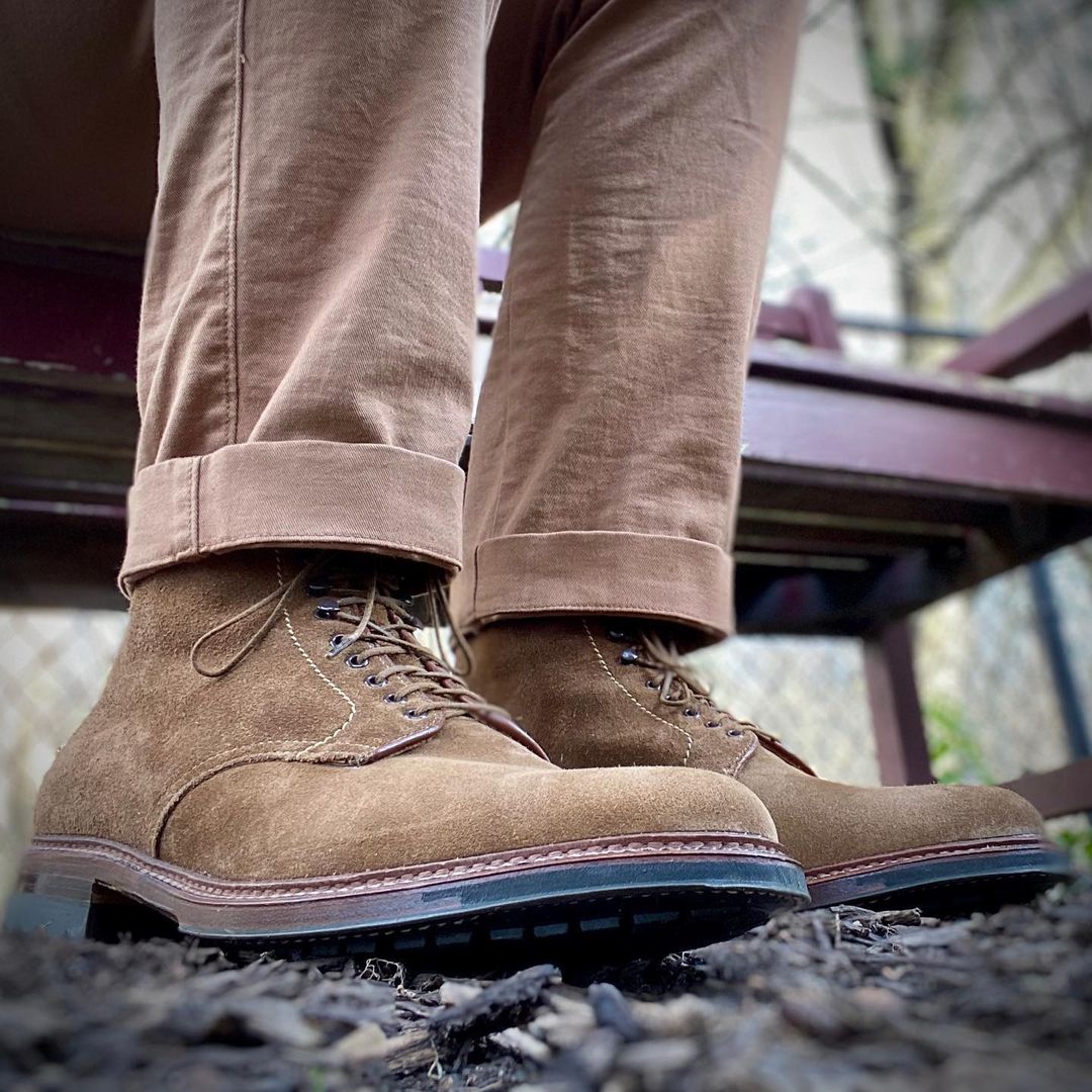 A person wears Alden Madisons 4511HC - Plain Toe Boot in Snuff Suede, crafted on a Barrie last with laces, paired with rolled-up tan pants. They stand on a wood chip-covered ground, the commando sole ready for adventure, against a blurred outdoor backdrop.