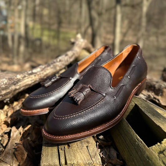 A pair of Alden Madisons D9120F Tassel Loafers in Brown Regina Grain sits on a wood surface outdoors, their Flex Leather Soles glistening in the sunlight against a gentle blur of trees and leaves.
