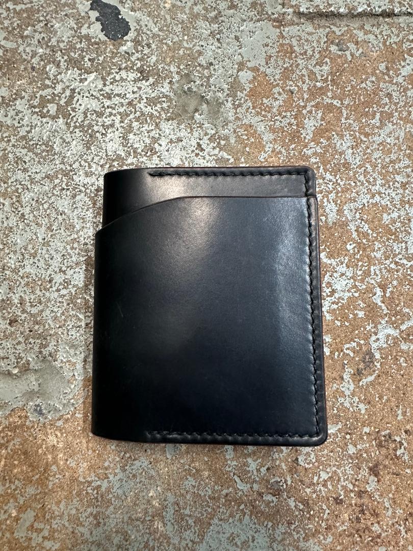 The Alden Madison Herb Bi-Fold Wallet in Shell Cordovan, handcrafted with visible stitching, lies on a worn, textured brown and white surface.