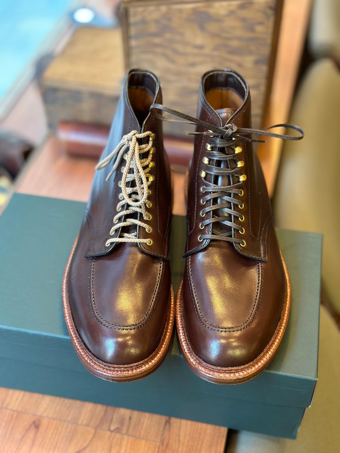 A pair of Alden Madisons 40577H Stitchdown Otis Indy Boots in brown calf rests atop a green shoebox. The boots, with contrasting tan and brown laces, sit on a wooden surface with a blurred background of other wooden items.