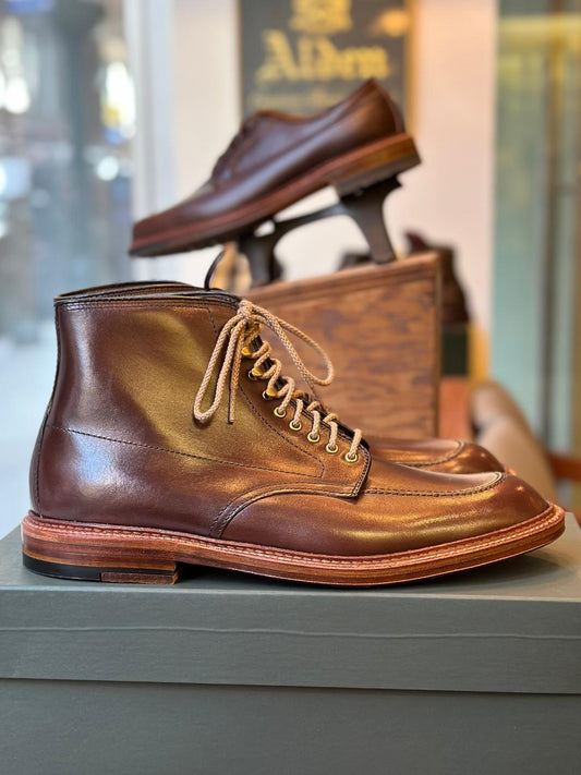 The 40577H - Stitchdown Otis Indy Boot in Brown Calf by Alden Madison shines on a green surface, with another pair on a wooden stand in the softly lit store, highlighting the allure of these brown calfskin lace-up boots with a double waterlock sole.