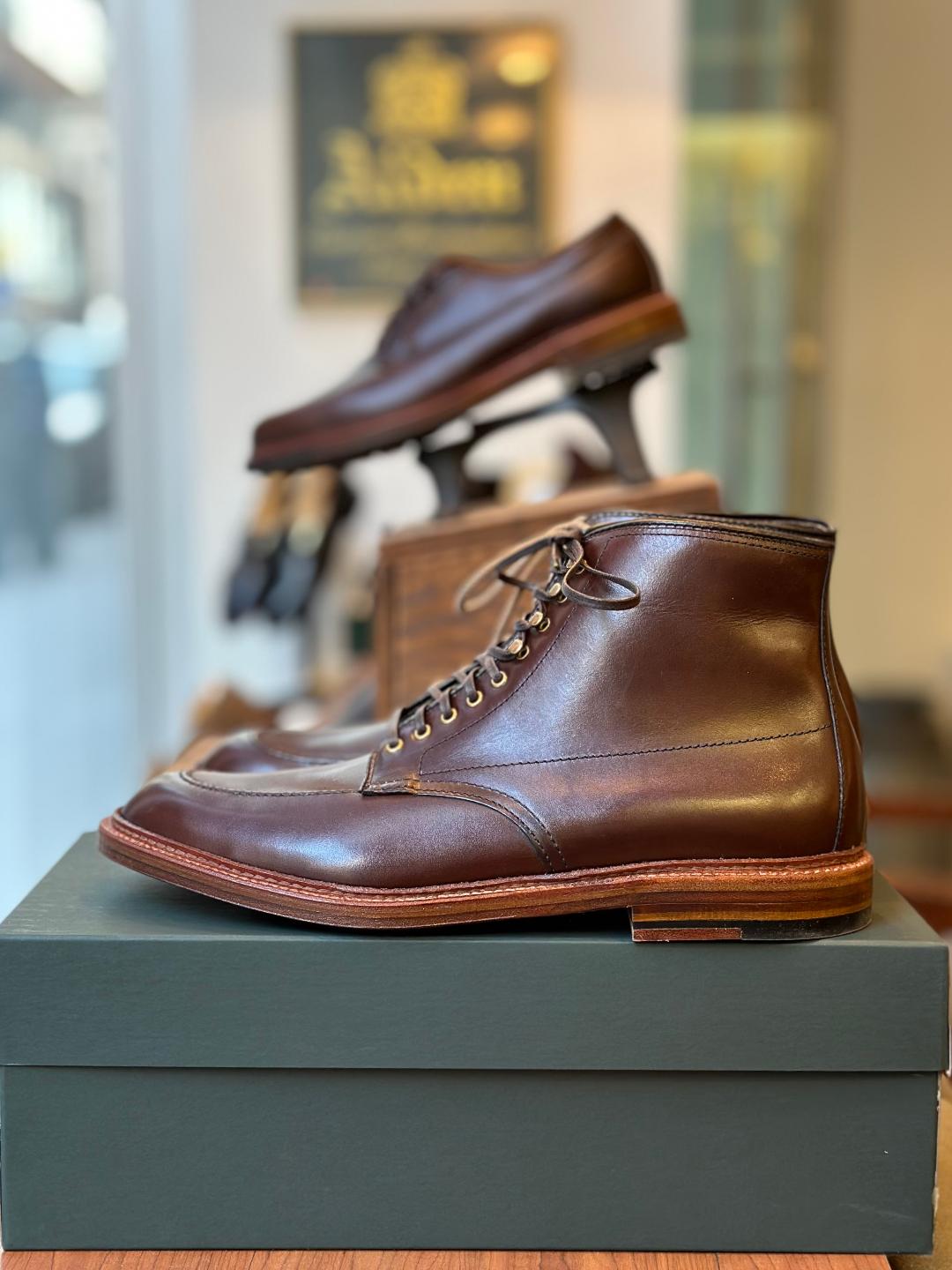 A close-up of the Alden Madison 40577H - Stitchdown Otis Indy Boot in Brown Calf with laces on a green shoebox. A low-cut shoe with a Double Waterlock Sole is elevated on a wooden stand in the background. Both shoes have polished finishes and are showcased in a store setting.