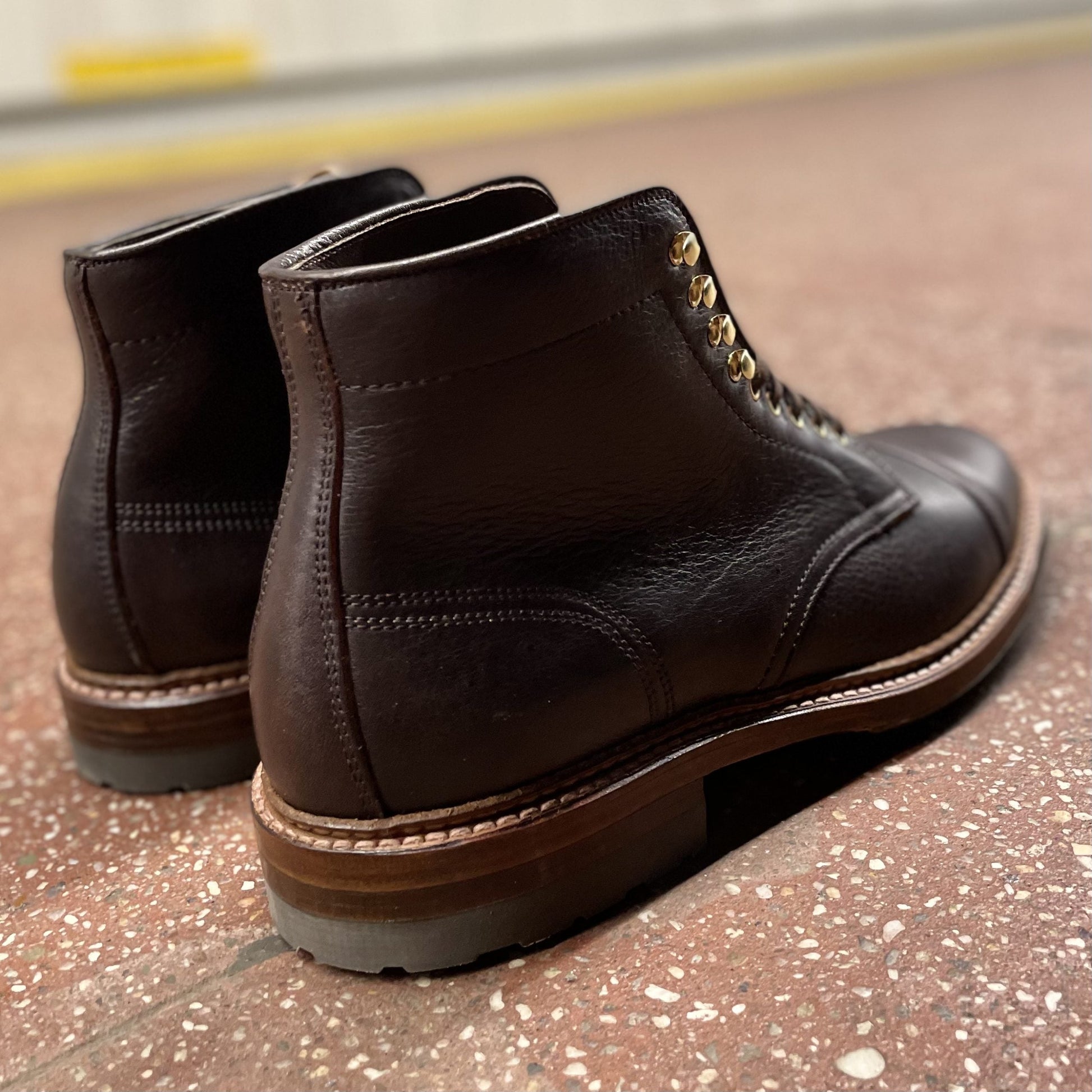 Alden Madison’s D1808HC - Stitchup 2/2 Boot in Arabica Lux7 calfskin, dark brown with gold eyelets and a sturdy Commando Sole, shown from the back and side on a reddish speckled floor.