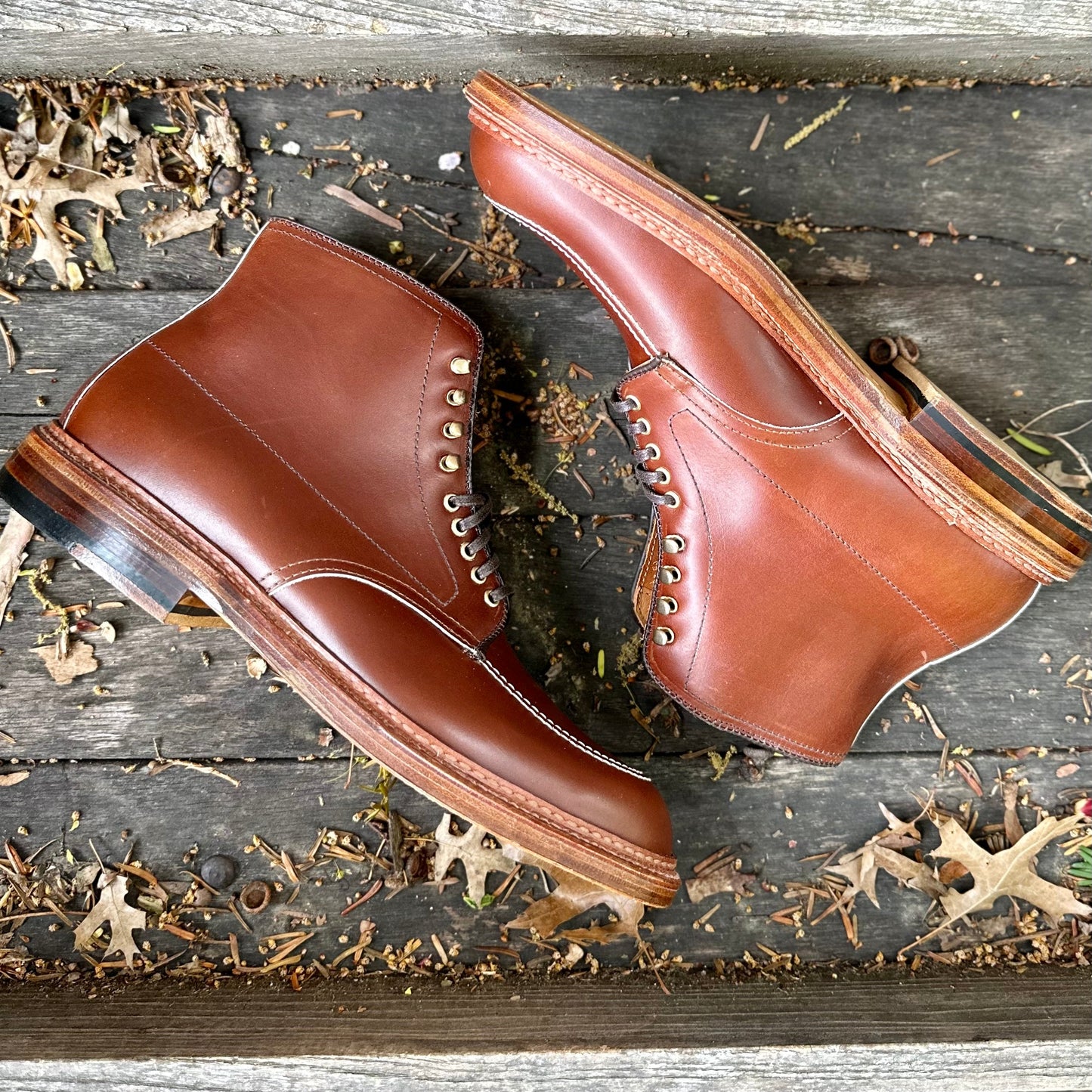 A pair of Alden Madisons D2940H - Madison Indy Boots in rich brown calf, featuring contrasting soles, sits on a wooden surface. Scattered dry leaves enhance the rustic ambiance, perfectly embodying the essence of the classic Indy Boot style.
