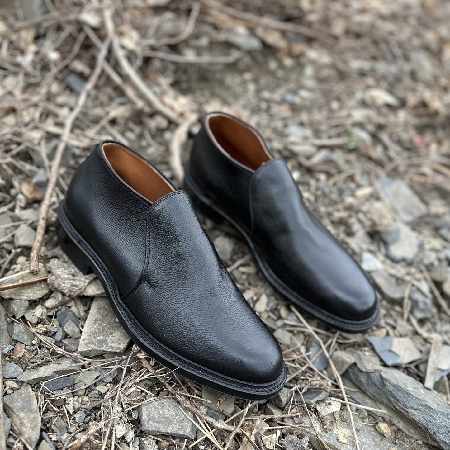 The Alden Madison D2711C - 750 Chukka Boot in Black Regina Grain with a textured finish and Commando rubber sole is placed on rocky, uneven ground with scattered dry twigs and stones.