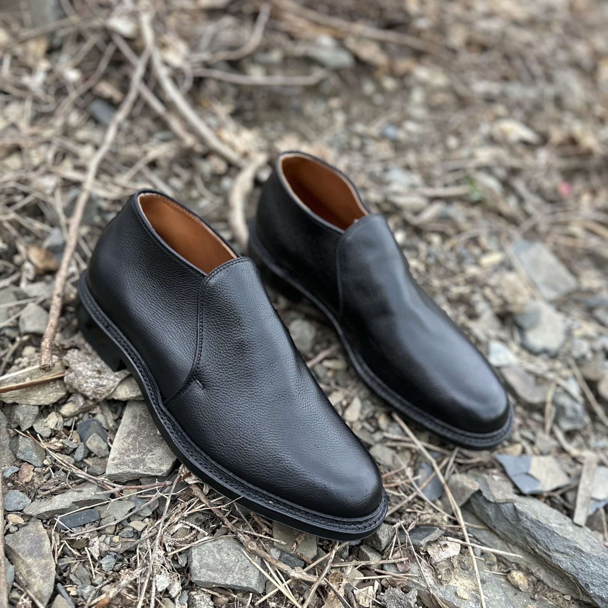 The Alden Madison D2711C - 750 Chukka Boot in Black Regina Grain with a textured finish and Commando rubber sole is placed on rocky, uneven ground with scattered dry twigs and stones.