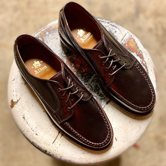 A pair of Alden Madison D9613C Ranger Mocc shoes in Color 8 Shell Cordovan with laces and antique edge, neatly set on a worn white round stool. The gold Alden logo shines inside the leather-lined interior.