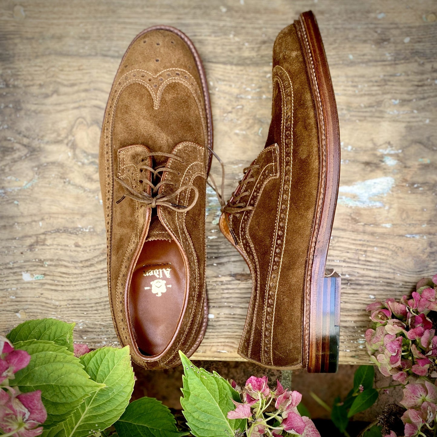 A pair of 9794 Long Wing Blucher in Snuff Suede by Alden Madison is displayed on a rustic wooden surface. The left shoe is upright, the right reveals the Waterlock leather sole, framed by pink and green flowers in the images lower corners.