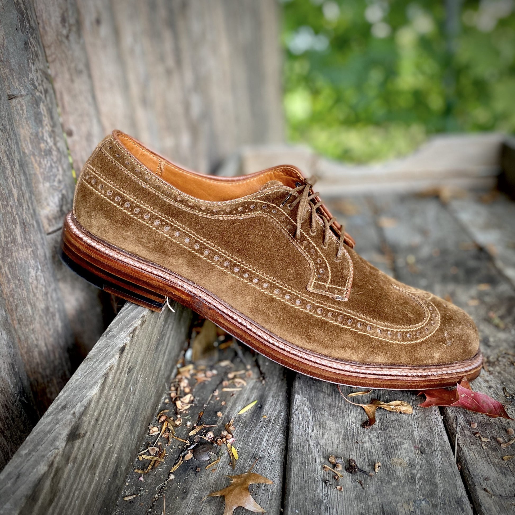 The Alden Madison 9794 - Long Wing Blucher in Snuff Suede, crafted on the Barrie last, sits on weathered wooden steps outdoors. Detailed stitching and blurred foliage emphasize its texture and craftsmanship, with small leaves scattered around.