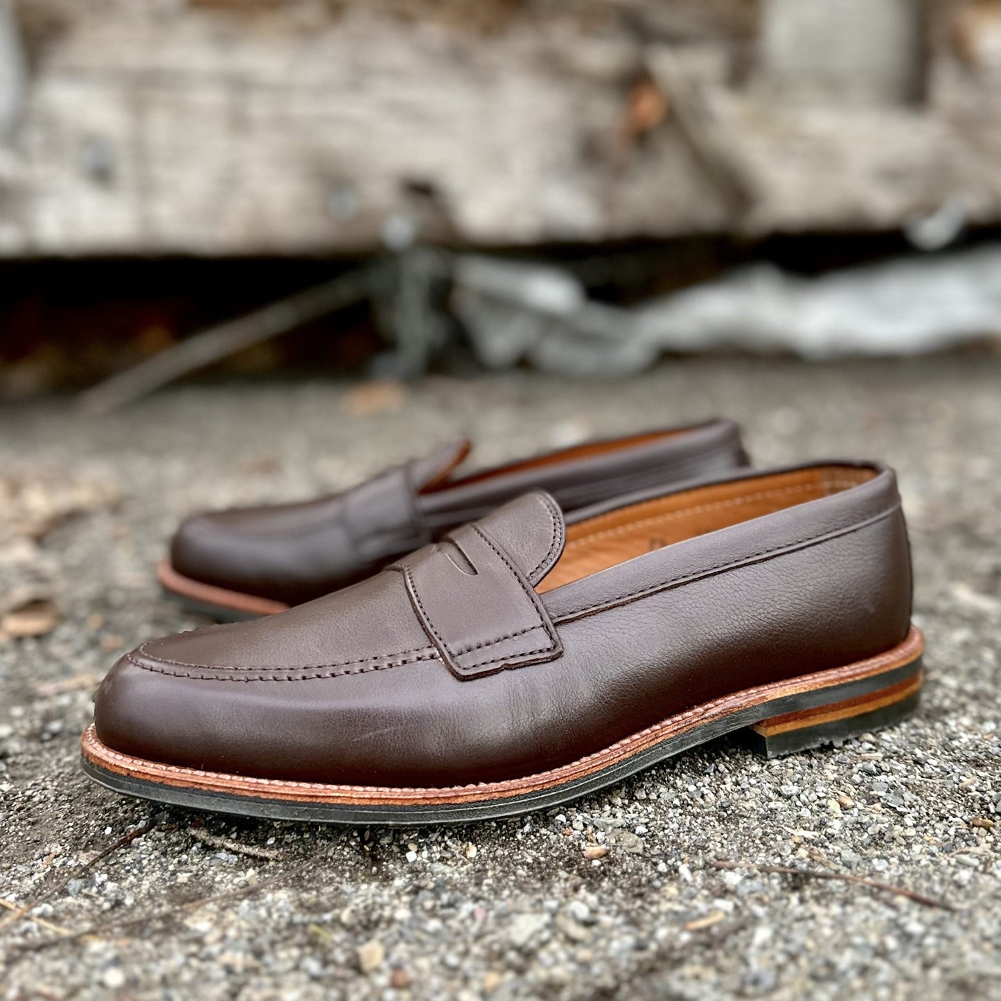 The Alden Madison D2203L Penny Loafer in Brown Lady Calf, featuring rubber soles, sits on a rough concrete surface with a slightly blurred wooden structure in the background.