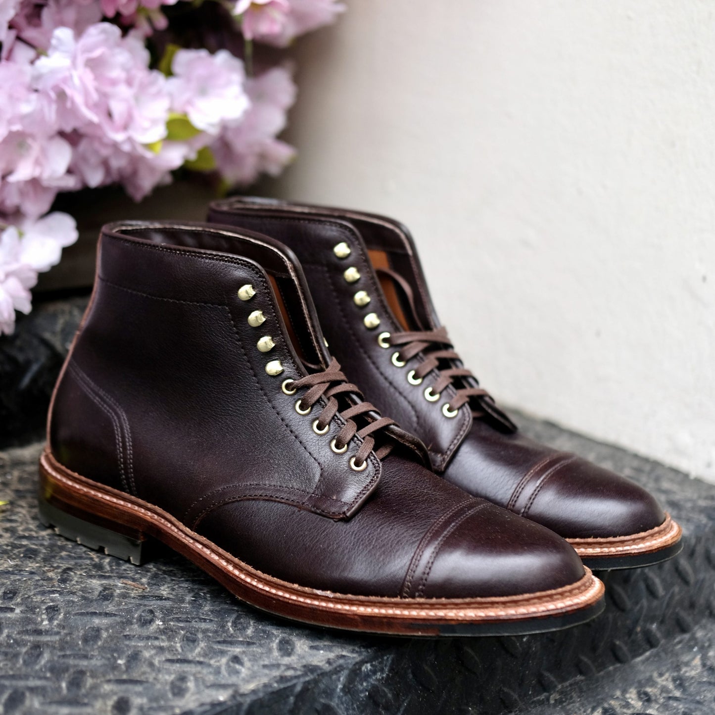 A pair of Alden Madison D1808HC - Stitchup 2/2 Boot in Arabica Lux7, featuring dark brown calfskin with brass eyelets and a stitched Commando Sole, displayed on a black surface near pink flowers and a light wall.