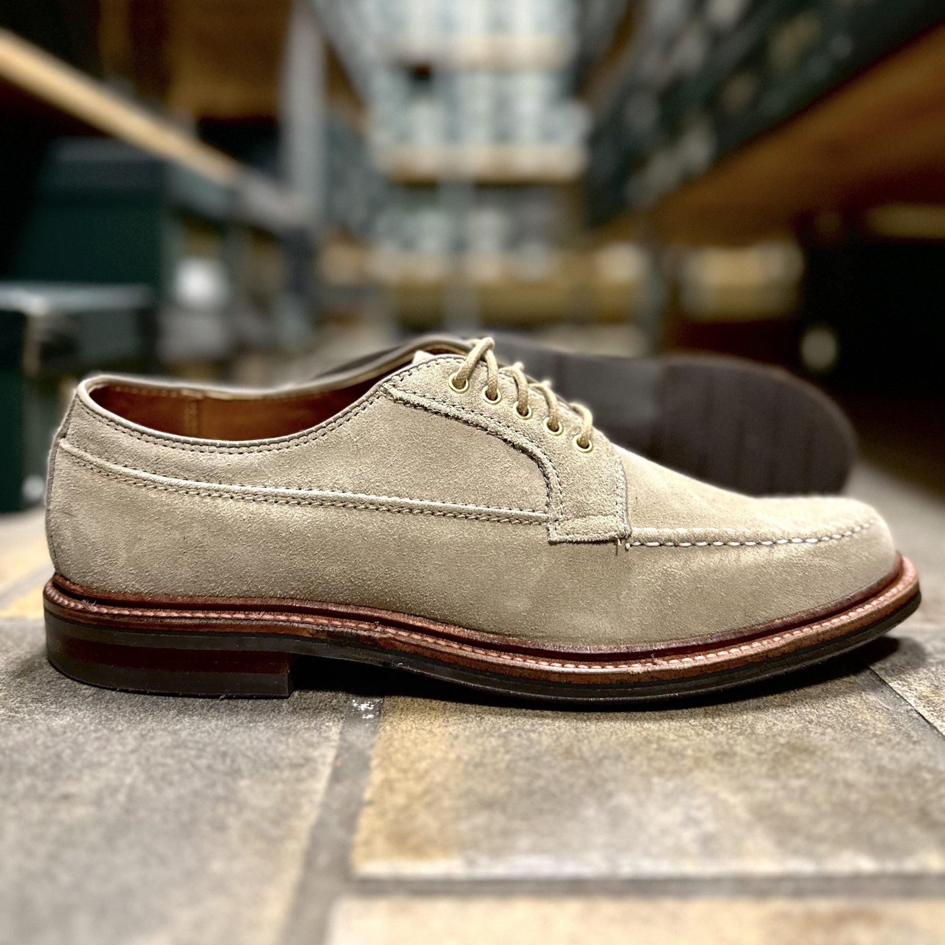 The Alden Madison D1634L - Ranger Mocc in Milkshake Suede, featuring brown laces and an Antique 360 Flat Welt, is displayed on a stone floor with blurred shelves of shoe boxes in the background.