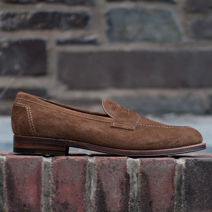 The Alden Madison 9693F - Flex Penny Loafer in Snuff Suede features stitching details, a low wooden heel, and a flex leather sole for comfort and style, shown resting on a brick ledge against a stone wall.