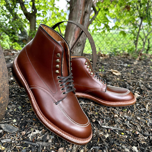 Pair of Alden Madisons D2940H - Madison Indy Boot is polished in brown calf leather with black laces, metal eyelets, and stitched sole. Displayed on dark soil with a lush background of green leaves and chain-link fence, these boots radiate timeless elegance.