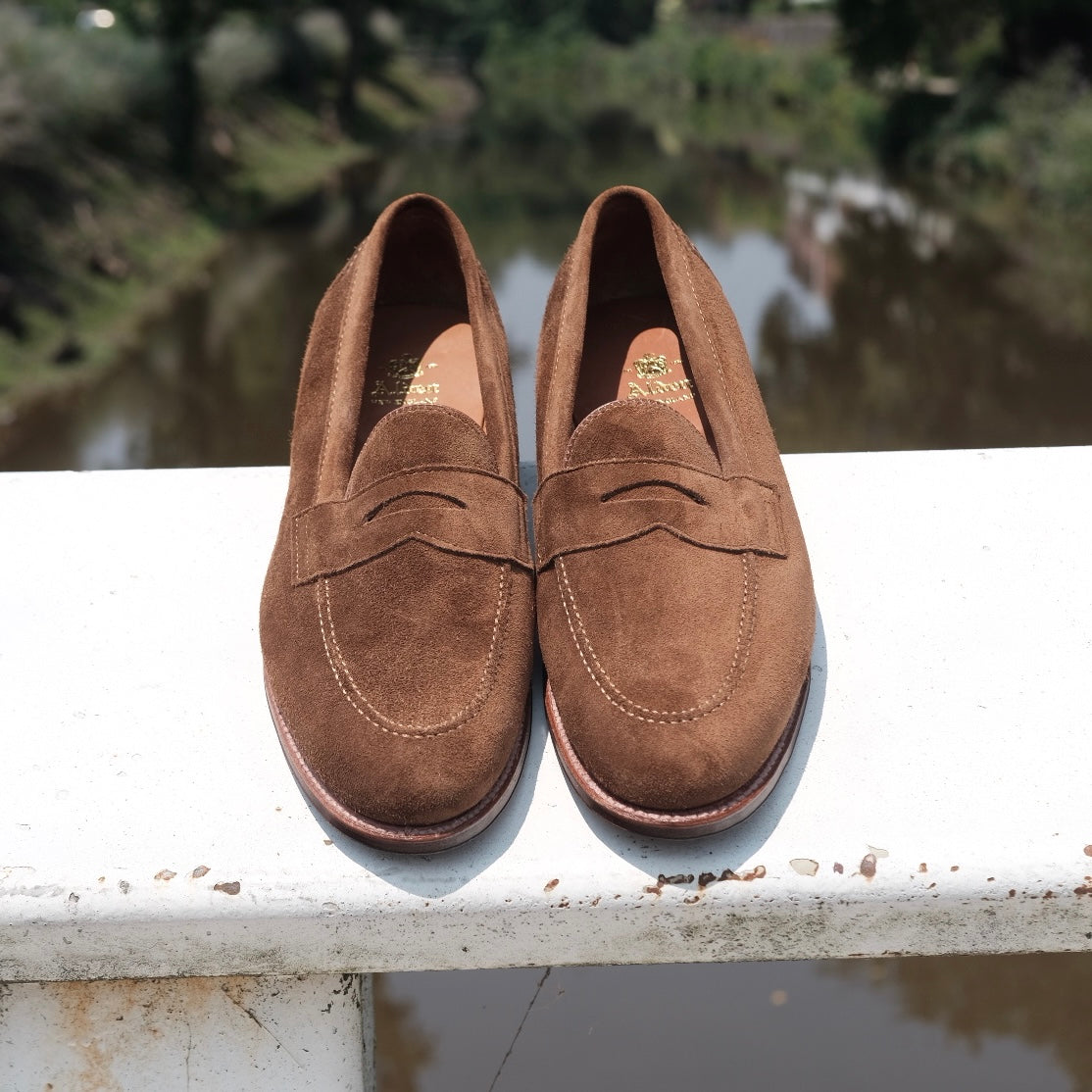 A pair of Alden Madison 9693F Flex Penny Loafers in snuff suede with Flex Leather Sole sits side by side on a white ledge, backed by a blurred scene of water and greenery.