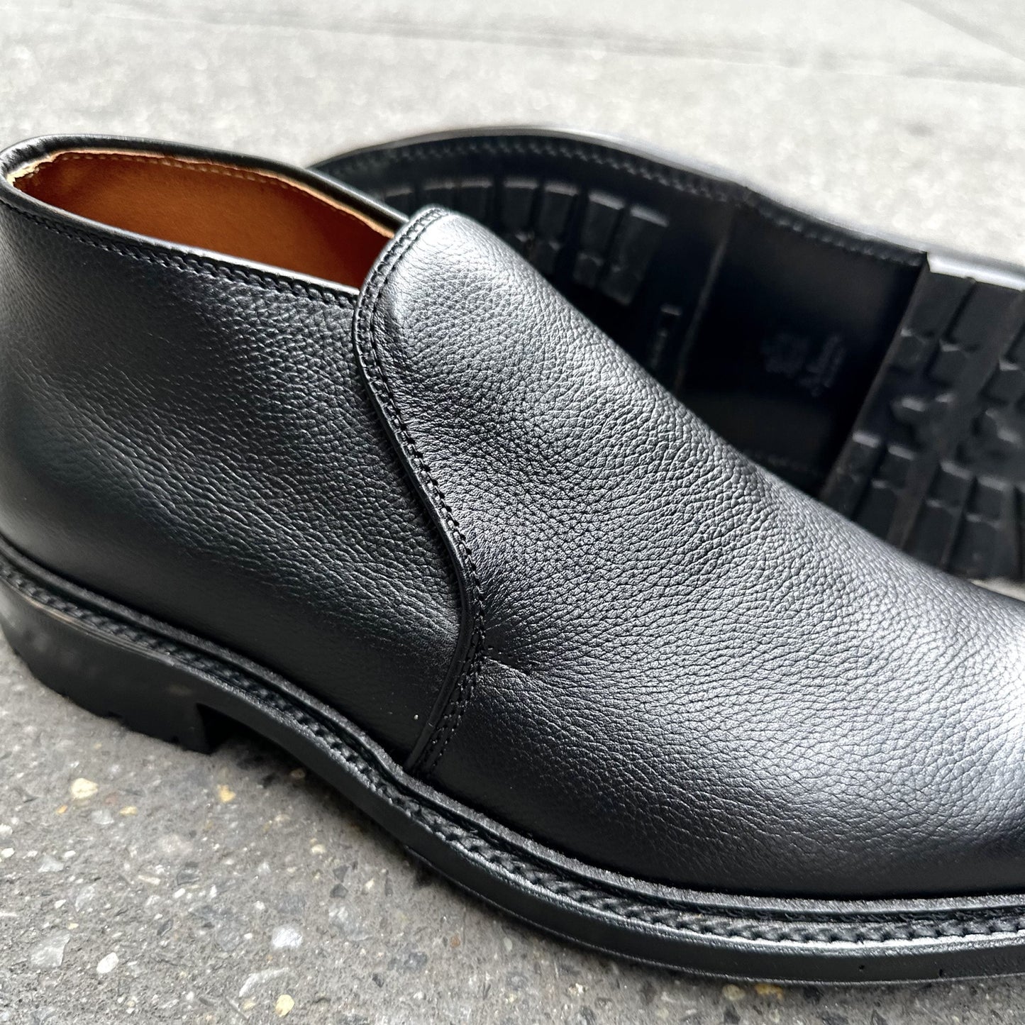 A close-up of the Alden Madison D2711C - 750 Chukka Boot in Black Regina Grain on concrete, with one upright and one on its side to showcase the textured leather and Commando Rubber Sole tread.