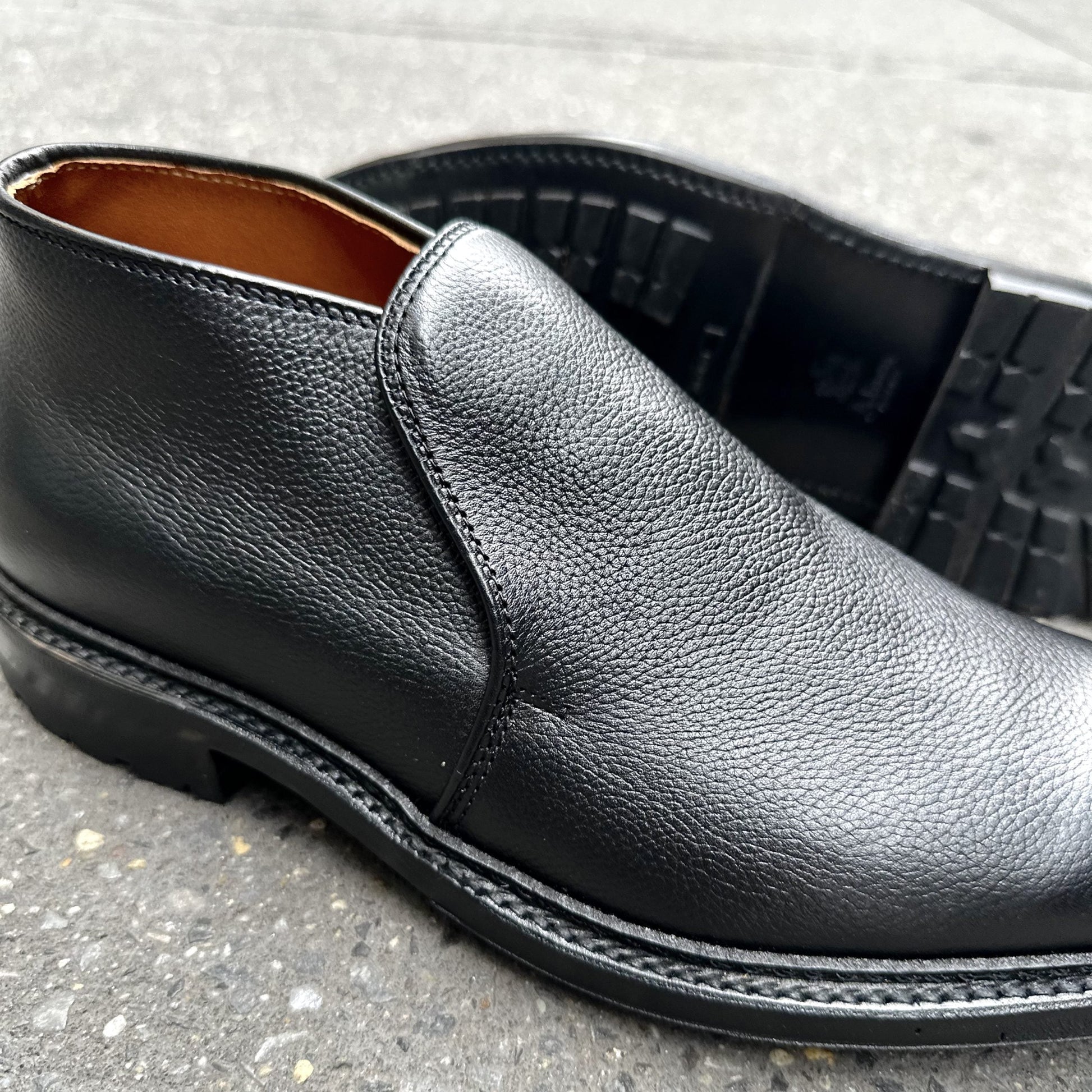 A close-up of the Alden Madison D2711C - 750 Chukka Boot in Black Regina Grain on concrete, with one upright and one on its side to showcase the textured leather and Commando Rubber Sole tread.