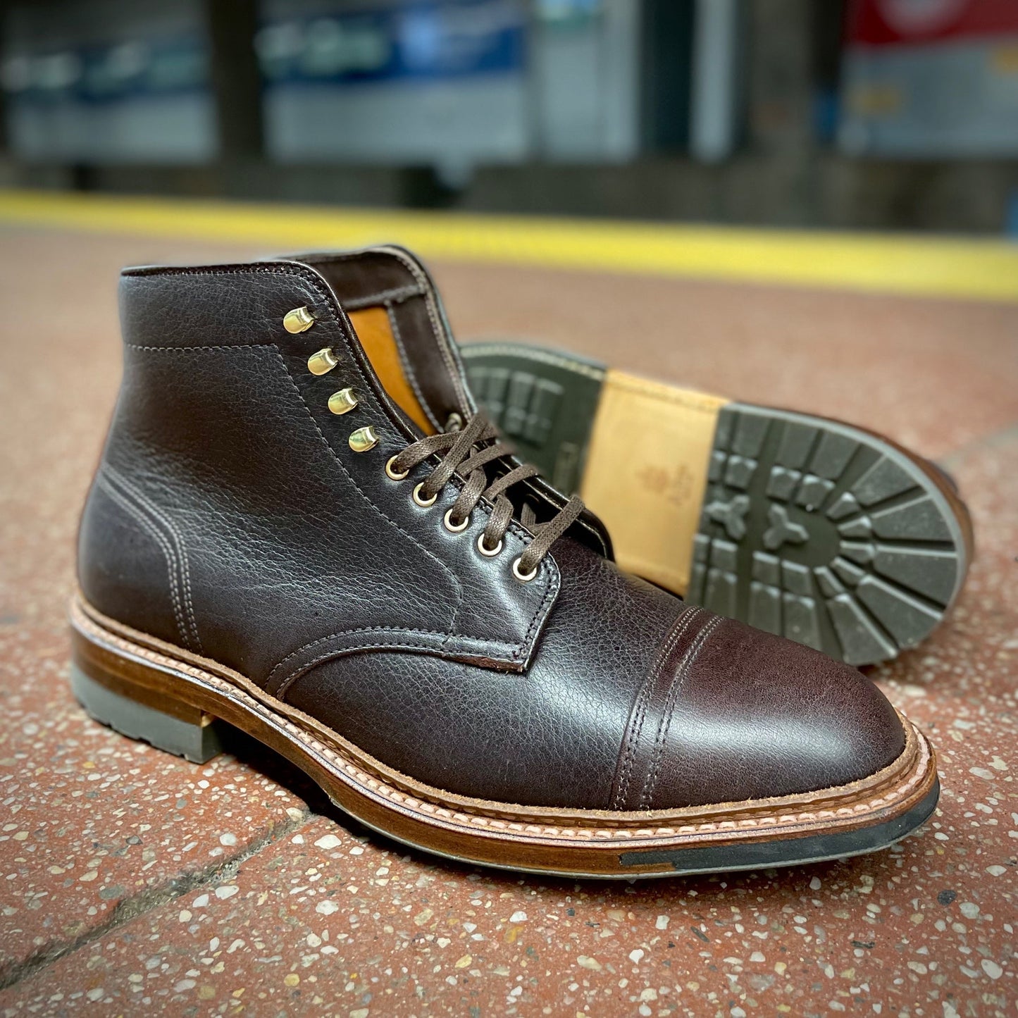 The Alden Madison D1808HC - Stitchup 2/2 Boot in Arabica Lux7, dark brown calfskin with cap toes, gold eyelets, and a sturdy Commando Sole, is displayed on a speckled floor, with one boot upright and one on its side.