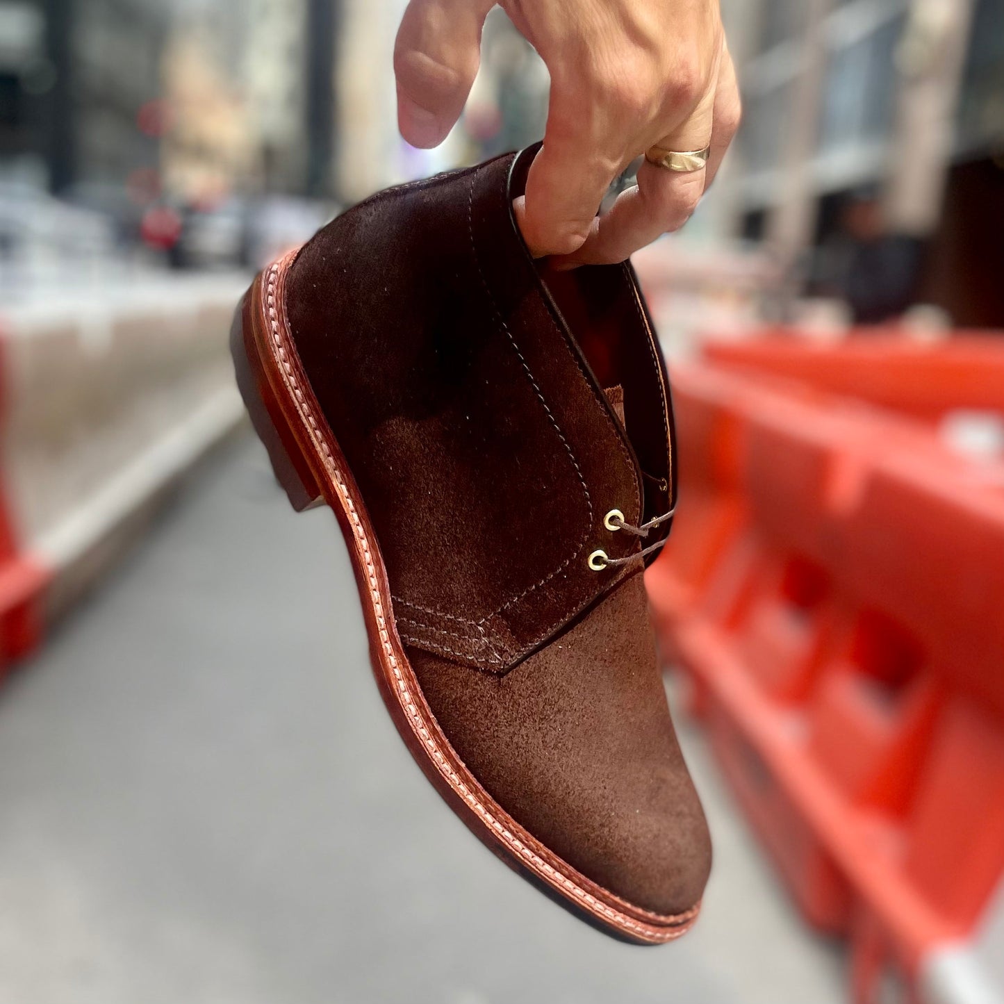 A hand holds the Alden Madison D1717C - Catskills Chukka Boot in Tobacco Reverse Chamois by the heel on a city street, with blurred orange construction barriers and buildings behind. The boot features a durable Commando sole for city adventures.