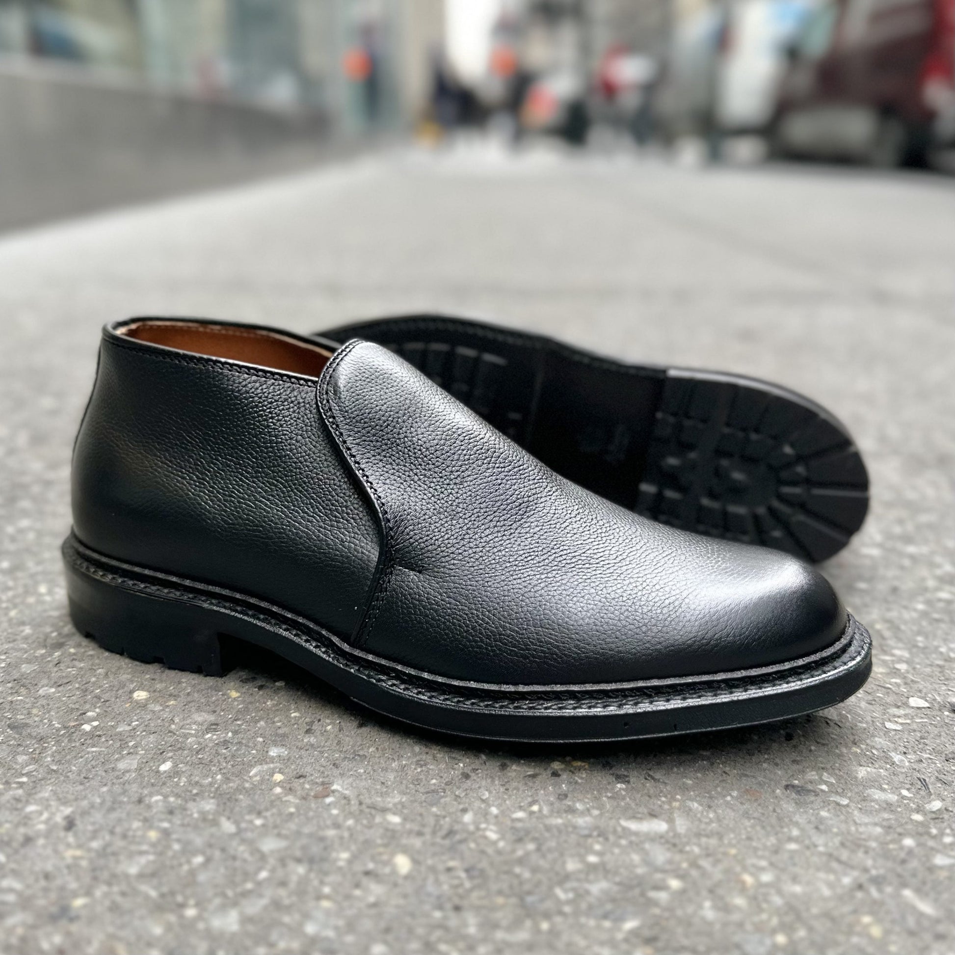A pair of Alden Madison D2711C - 750 Chukka Boots in Black Regina Grain leather with a Commando rubber sole is displayed on a city sidewalk—one upright, the other on its side showing the tread. The background appears slightly blurred.